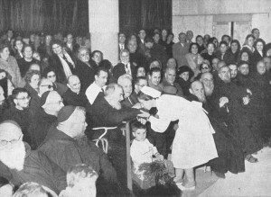 Padre Pio at the Home for the Relief of Suffering attends a program in the auditorium. A staff nurse greets Padre Pio and kisses his hand.