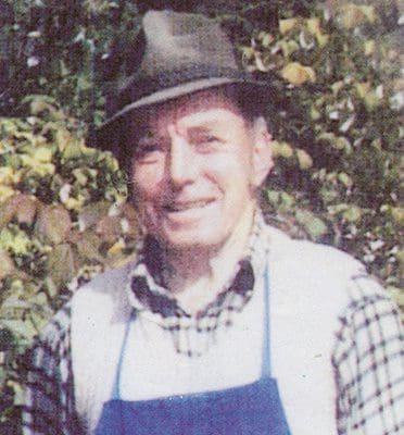 A man wearing an apron and hat standing in front of bushes.