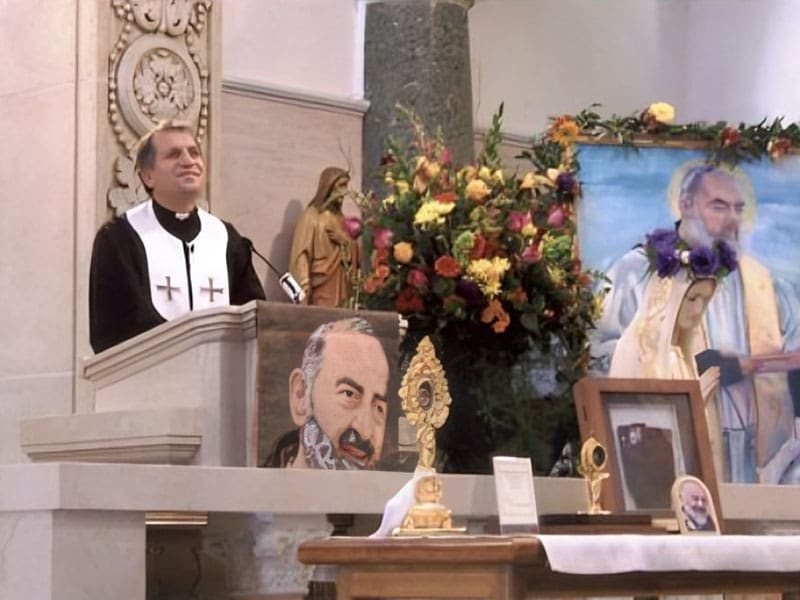 A priest is standing in front of some paintings.