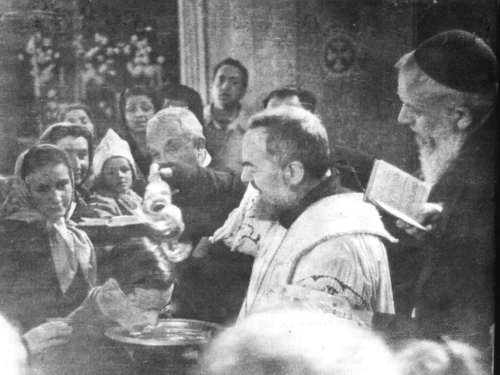 Priest baptizing child surrounded by onlookers.