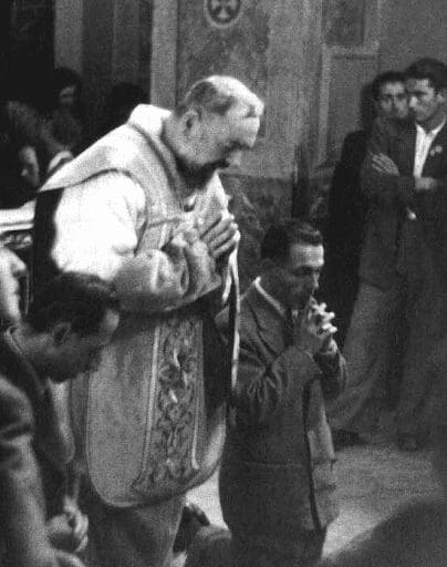 A clergyman in ceremonial robes interacting with a kneeling man in a crowd.