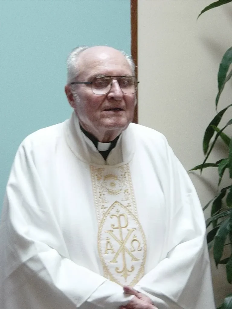Elderly priest in white robe, indoors.